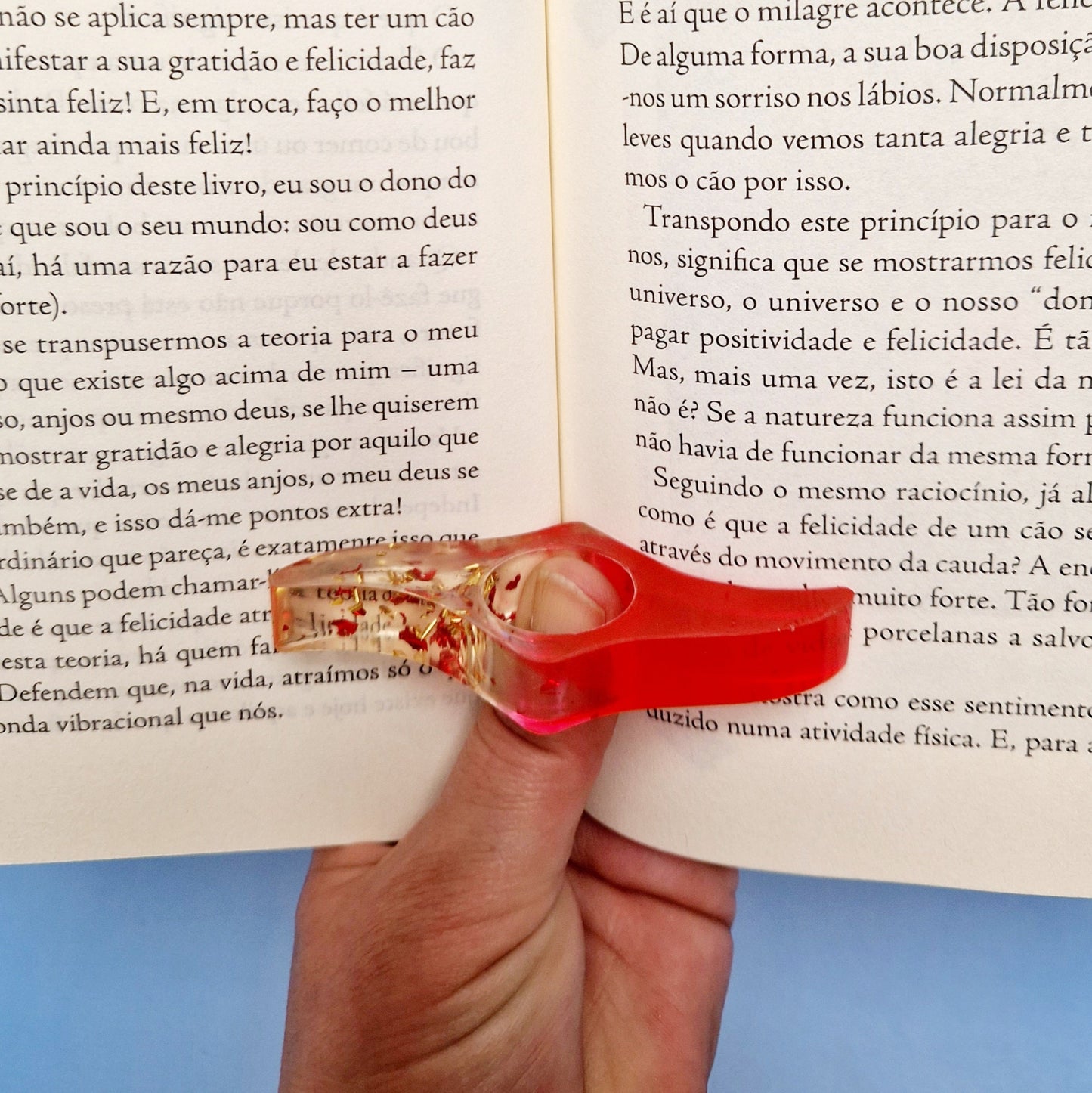 Anel de leitura em resina vermelha translúcida com glitter dourado, encaixado no centro do livro para facilitar a leitura com uma mão.