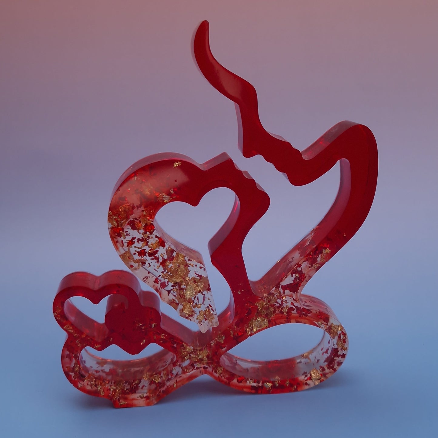 Escultura decorativa de casal em resina epóxi vermelha, com silhuetas abstratas formando um coração, acabamento translúcido e detalhes em folhas metálicas douradas, sobre fundo suave.