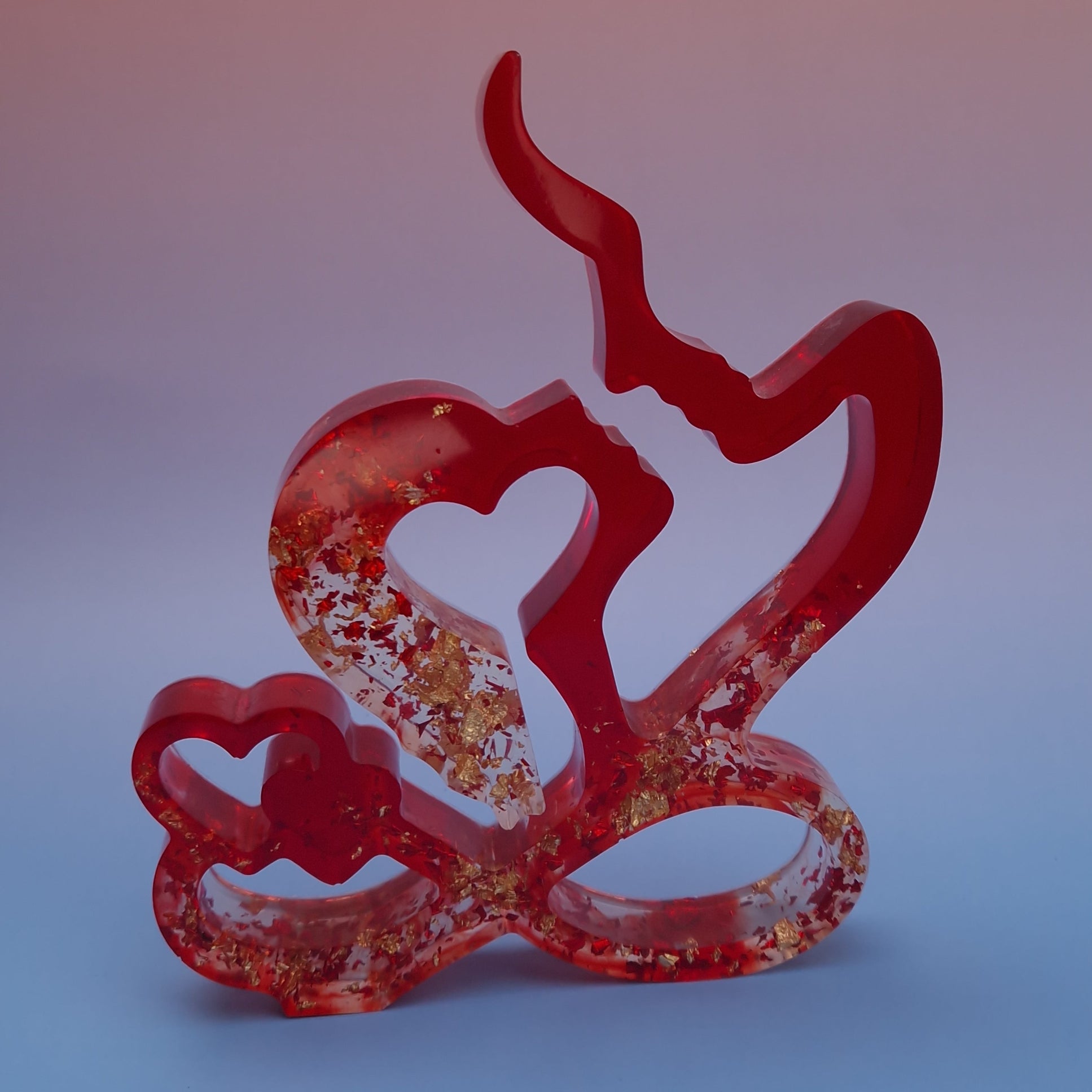 Escultura decorativa de casal em resina epóxi vermelha, com silhuetas abstratas formando um coração, acabamento translúcido e detalhes em folhas metálicas douradas, sobre fundo suave.