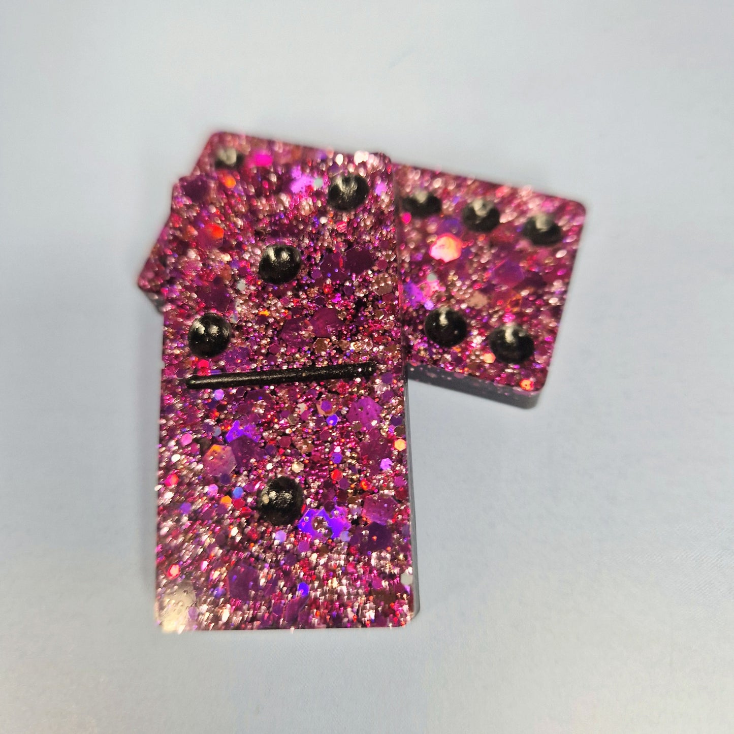 Duas peças de dominó artesanal em resina rosa com glitter intenso, sobrepostas, com detalhe dos pontos em relevo.