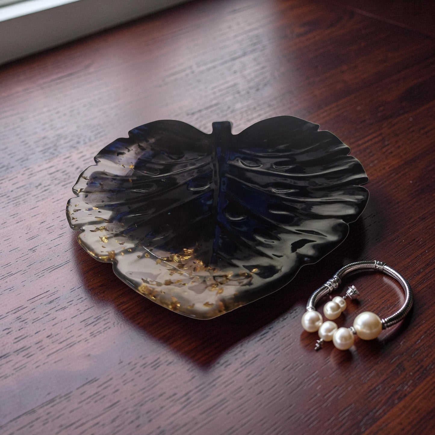 Bandeja de resina preta e ouro em formato de folha de Monstera com pulseira e brincos de pérola sobre mesa de madeira.